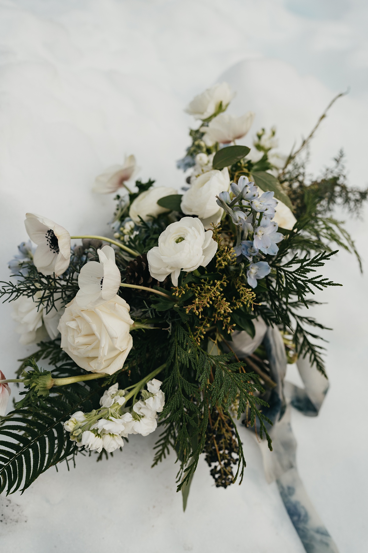 A white bouquet with evergreens and pops of blue flowers, sitting in the snow