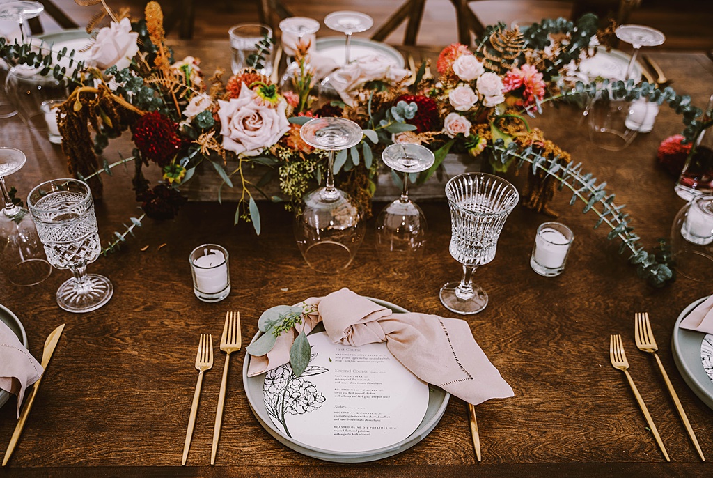 Bloom-Poet-Seattle-Wedding-Florist-Summer-Wedding-Farm-Tables.jpg A long farm table is set with a large floral arrangement, flatware, plates, napkins, and botanical sprigs